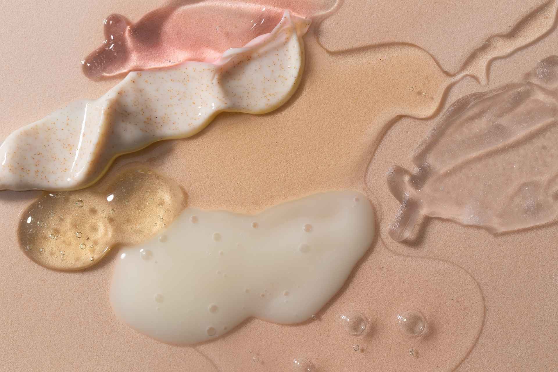Mehrere Hautpflege-Texturen wie Serum, Gel und Creme in verschiedenen Farben und Konsistenzen, nebeneinander auf einer beigefarbenen Oberfläche aufgetragen.