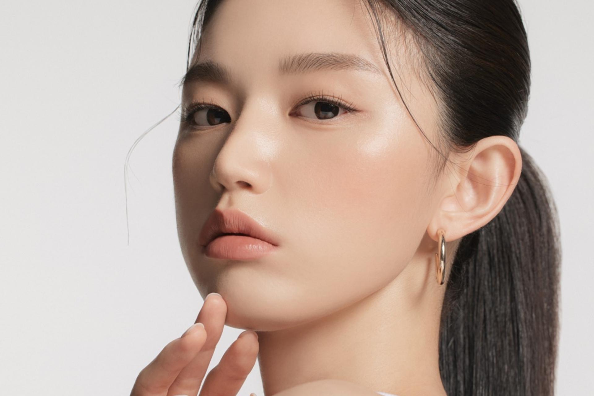 Nahaufnahme eines Beauty-Porträts mit makellos glowy Haut, natürlichem Make-up, goldenen Creolen und zurückgebundenem Haar; Fokus auf einem frischen, minimalistischen Look.